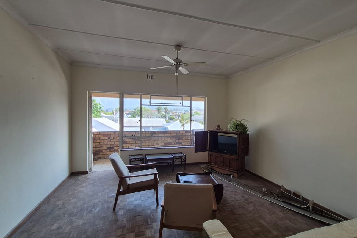 Spacious living room featuring parquet flooring, a ceiling fan, and large windows with access to a balcony, offering potential for modernization.