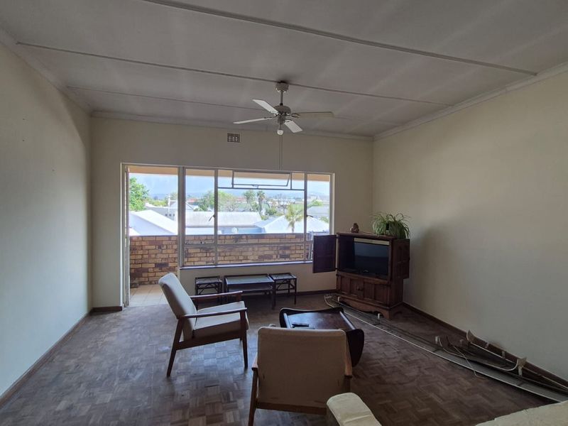 Spacious living room featuring parquet flooring, a ceiling fan, and large windows with access to a balcony, offering potential for modernization.