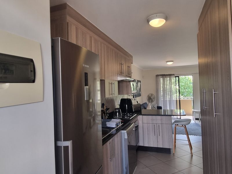 Open plan kitchen with modern wood-finish cabinetry and tiled flooring, leading through to a living area with access to a balcony.
