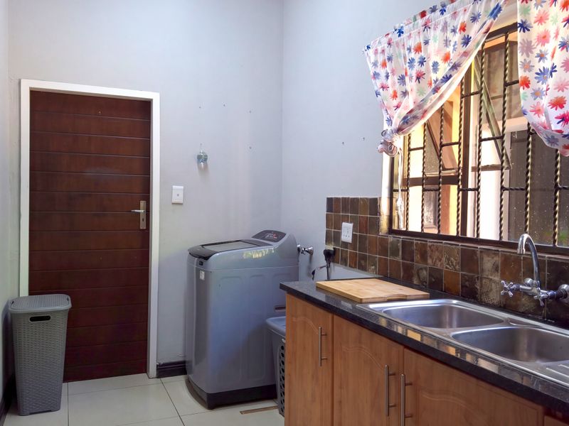 Functional laundry area featuring a double stainless steel sink, granite countertops, wooden cabinetry, and tiled splashback with security bars on the window.