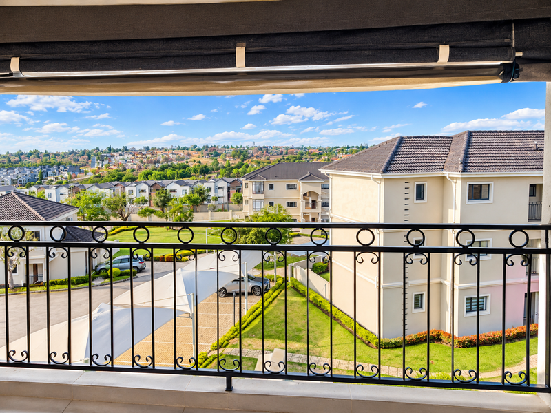 A spacious balcony featuring a decorative wrought iron balustrade, retractable awning, and elevated views over the surrounding residential complex and landscaped grounds.