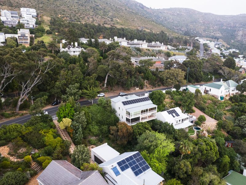 Aerial view of a residential property featuring solar panels on the roof, surrounded by lush greenery and established trees in a hillside neighborhood.