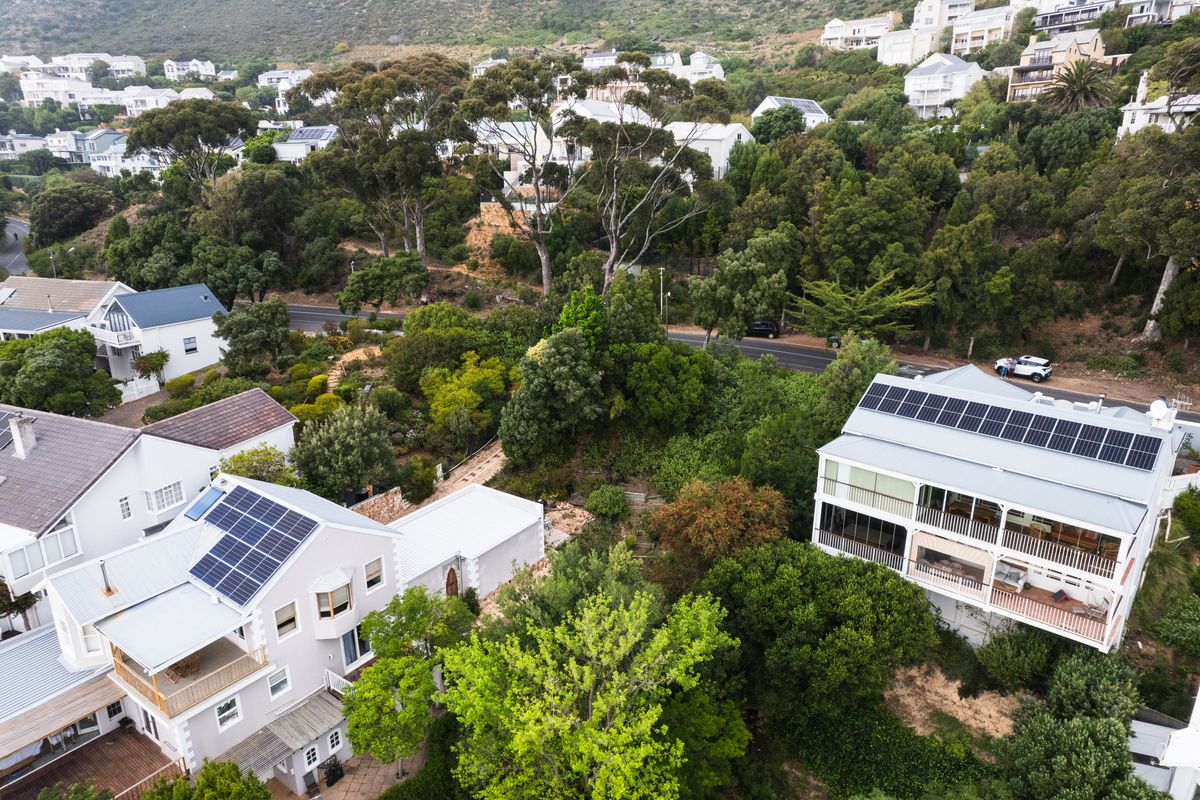 Aerial view of a residential property featuring multiple buildings with solar panel installations, surrounded by lush greenery and established trees in a hillside neighborhood.