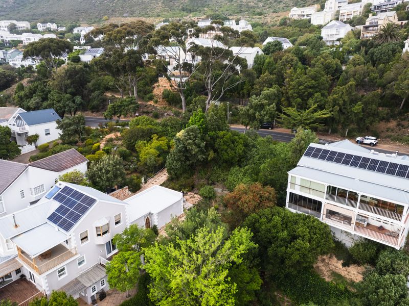 Aerial view of a residential property featuring multiple buildings with solar panel installations, surrounded by lush greenery and established trees in a hillside neighborhood.