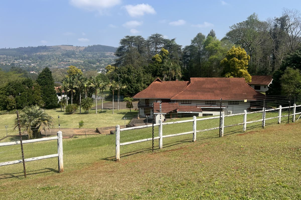 A wide-angle view of a residential property featuring a large lawn, mature trees, and a house with a tiled roof, secured by electric fencing.