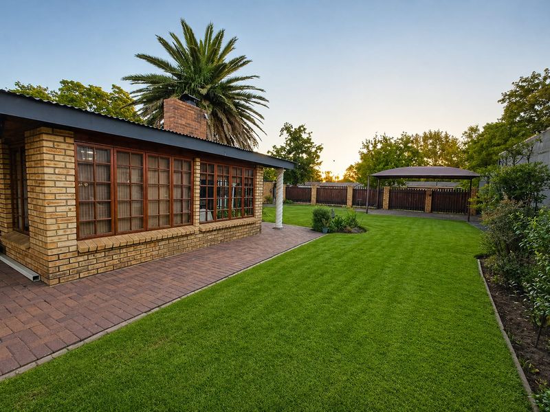A well-maintained rear garden featuring a paved patio area, brick exterior, timber-framed windows, and a detached carport set within a landscaped lawn.