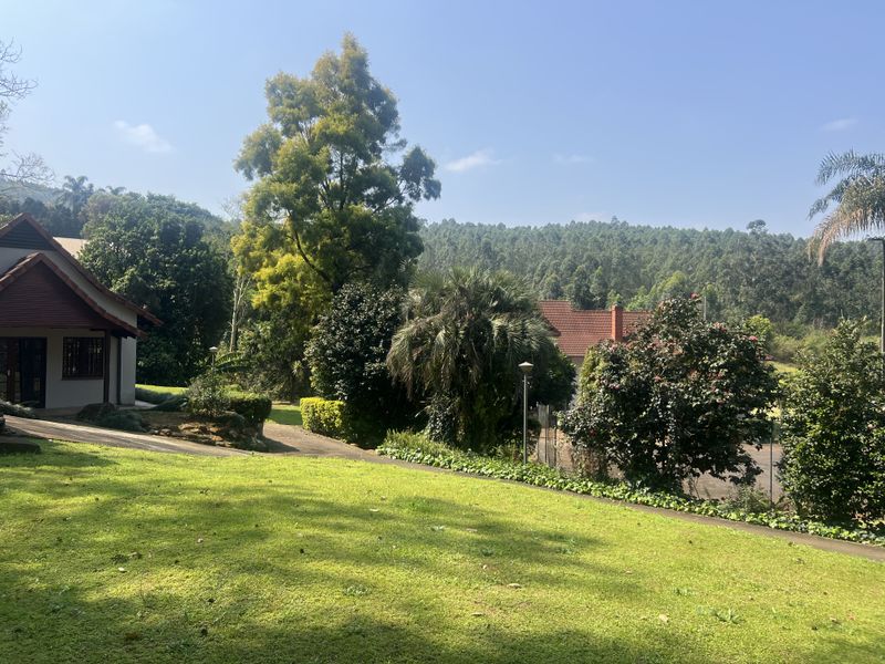 A well-maintained garden area featuring a lush green lawn, mature trees, and paved pathways, offering a serene outdoor setting with scenic hillside views.