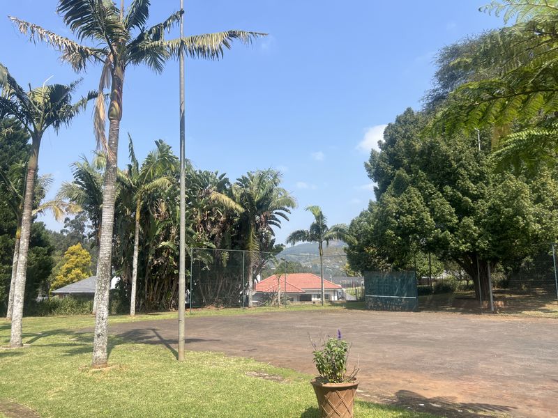 A private outdoor tennis court surrounded by mature trees and landscaped garden areas, featuring perimeter chain-link fencing.