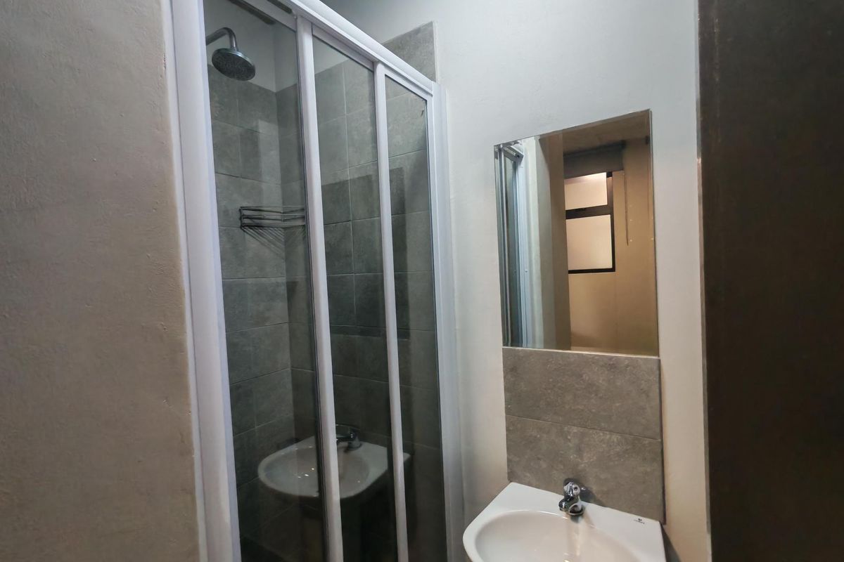 A compact, modern bathroom featuring a glass-enclosed shower, pedestal basin with mixer tap, and a wall-mounted mirror with tiled splashback.