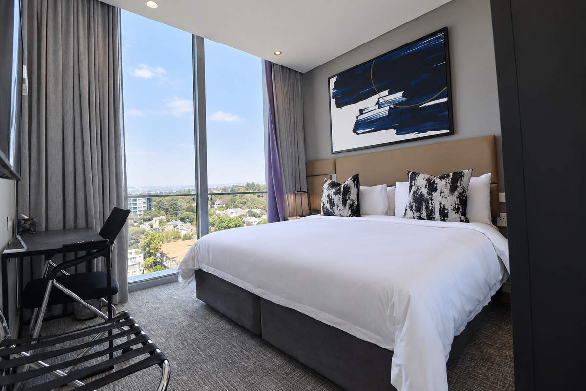 Modern bedroom featuring floor-to-ceiling windows with expansive views, neutral carpeting, and a built-in headboard in a well-maintained penthouse setting.