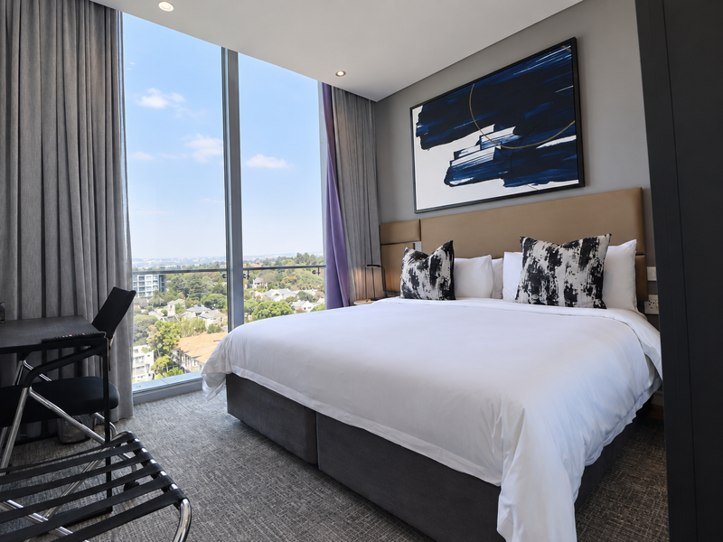 Modern bedroom featuring floor-to-ceiling windows with expansive views, neutral carpeting, and a built-in headboard in a well-maintained penthouse setting.