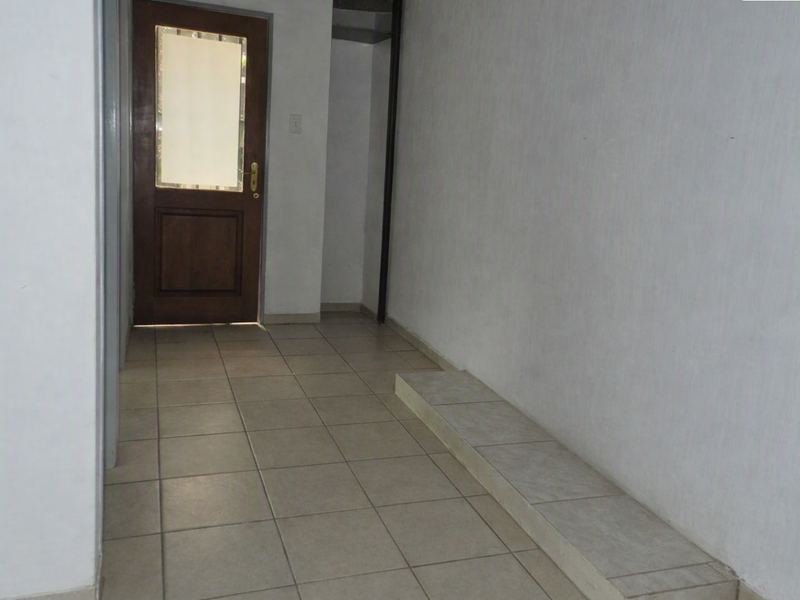 A simple, empty hallway featuring tiled flooring, a raised tiled platform, and a timber-framed entrance door with a glass panel.