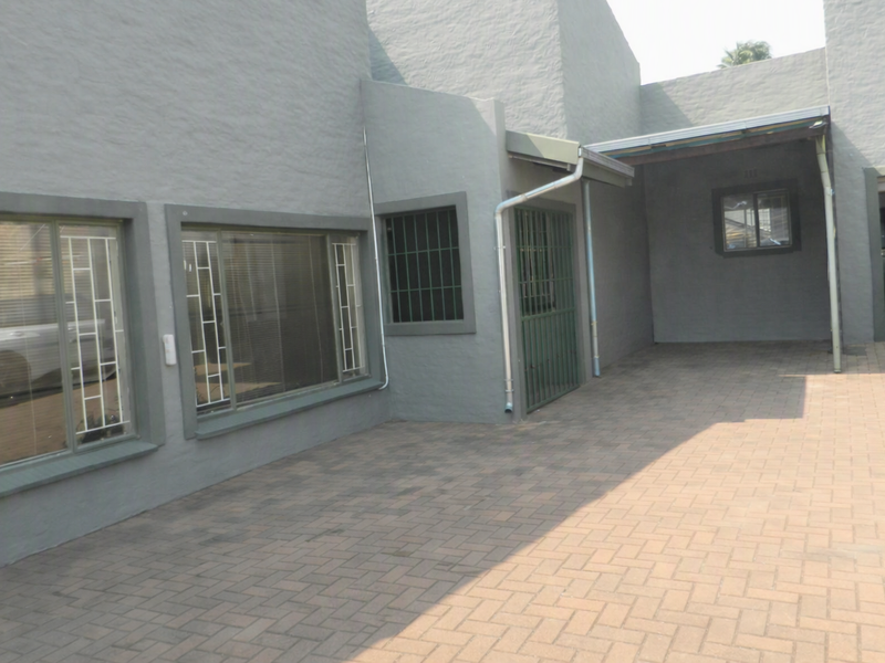 A paved driveway area leading to a covered carport and building entrance, featuring grey exterior walls and security bars on windows.