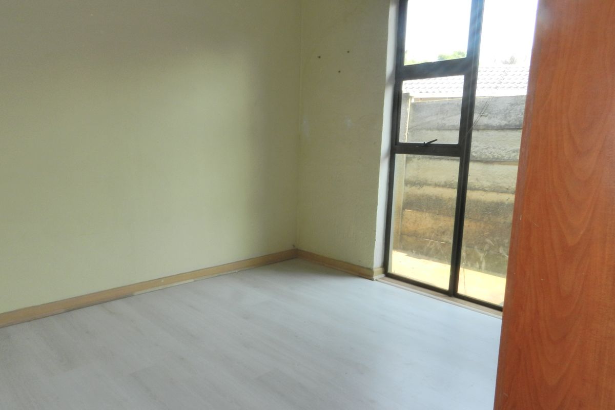 An empty bedroom featuring light-toned laminate flooring, neutral walls, and a large aluminium-framed window providing natural light.