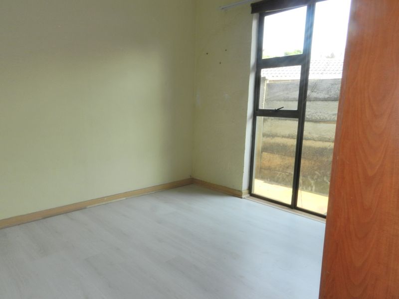 An empty bedroom featuring light-toned laminate flooring, neutral walls, and a large aluminium-framed window providing natural light.