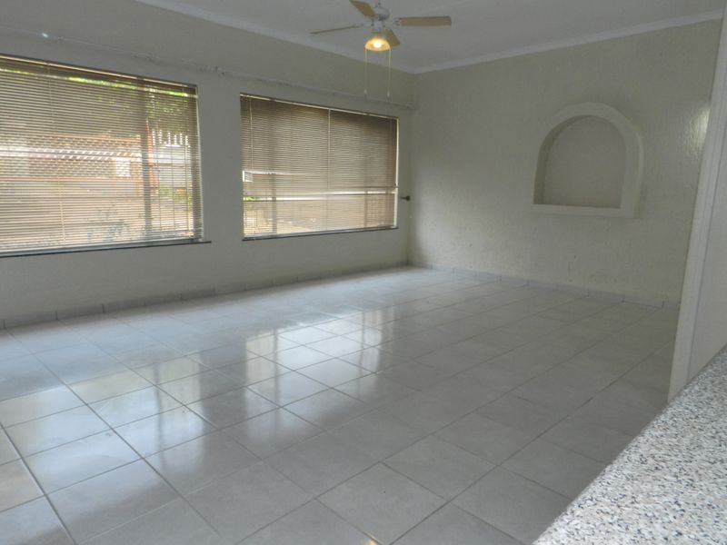 A spacious, empty living room featuring tiled flooring, a ceiling fan, and a built-in wall niche, illuminated by natural light through venetian blinds.