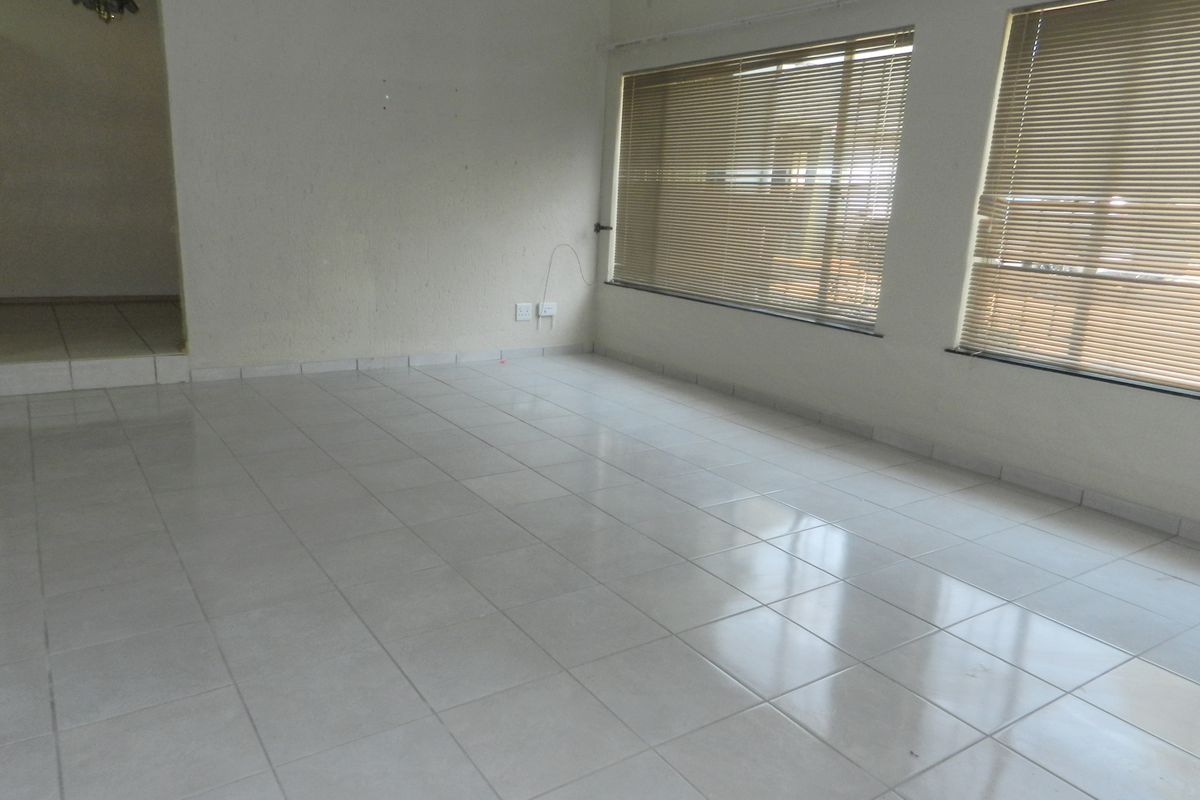 A bright, empty living room featuring neutral tiled flooring, large windows with fitted venetian blinds, and clean white walls.