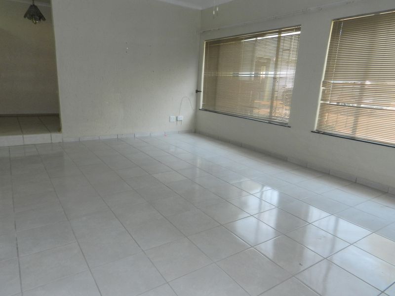 A bright, empty living room featuring neutral tiled flooring, large windows with fitted venetian blinds, and clean white walls.