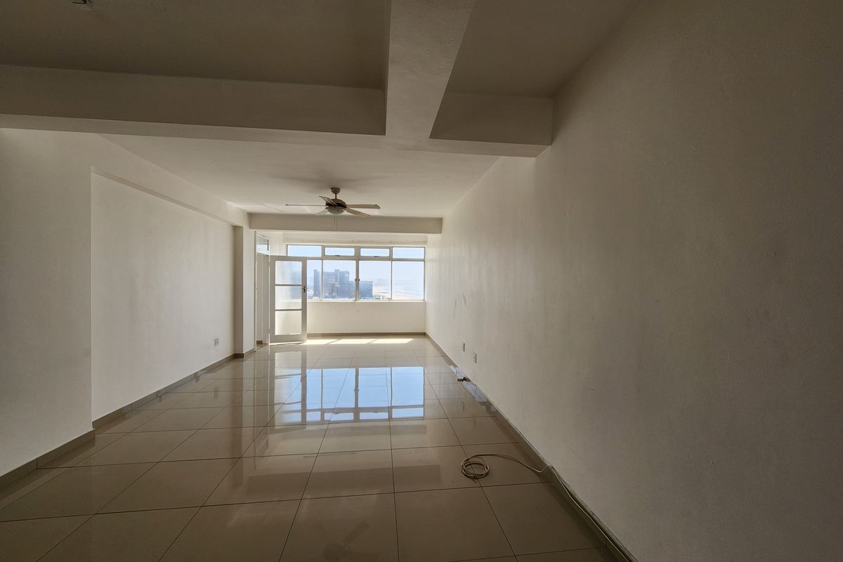 A spacious, empty living area featuring neutral tiled flooring, a ceiling fan, and large windows that provide natural light and an urban view.