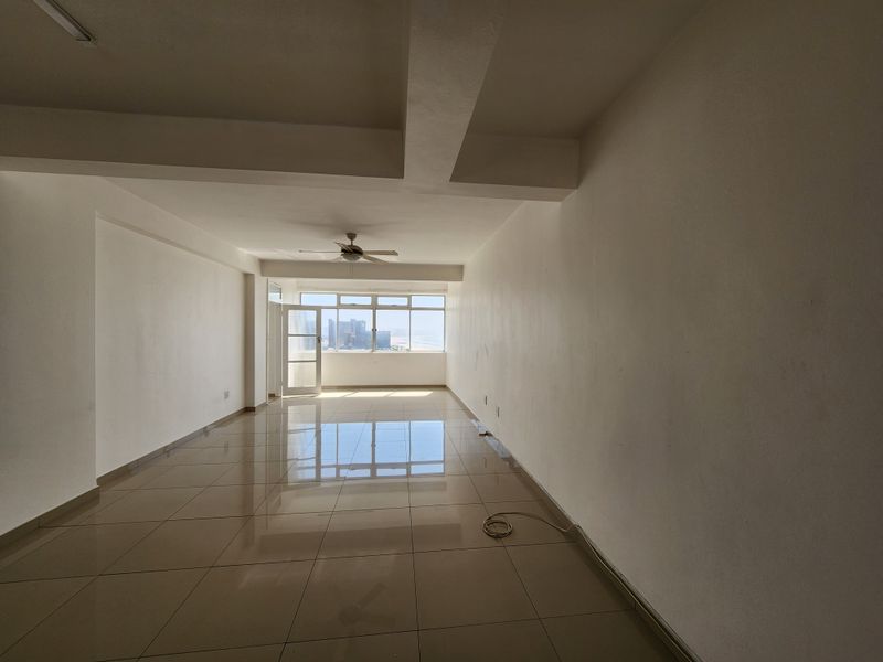 A spacious, empty living area featuring neutral tiled flooring, a ceiling fan, and large windows that provide natural light and an urban view.