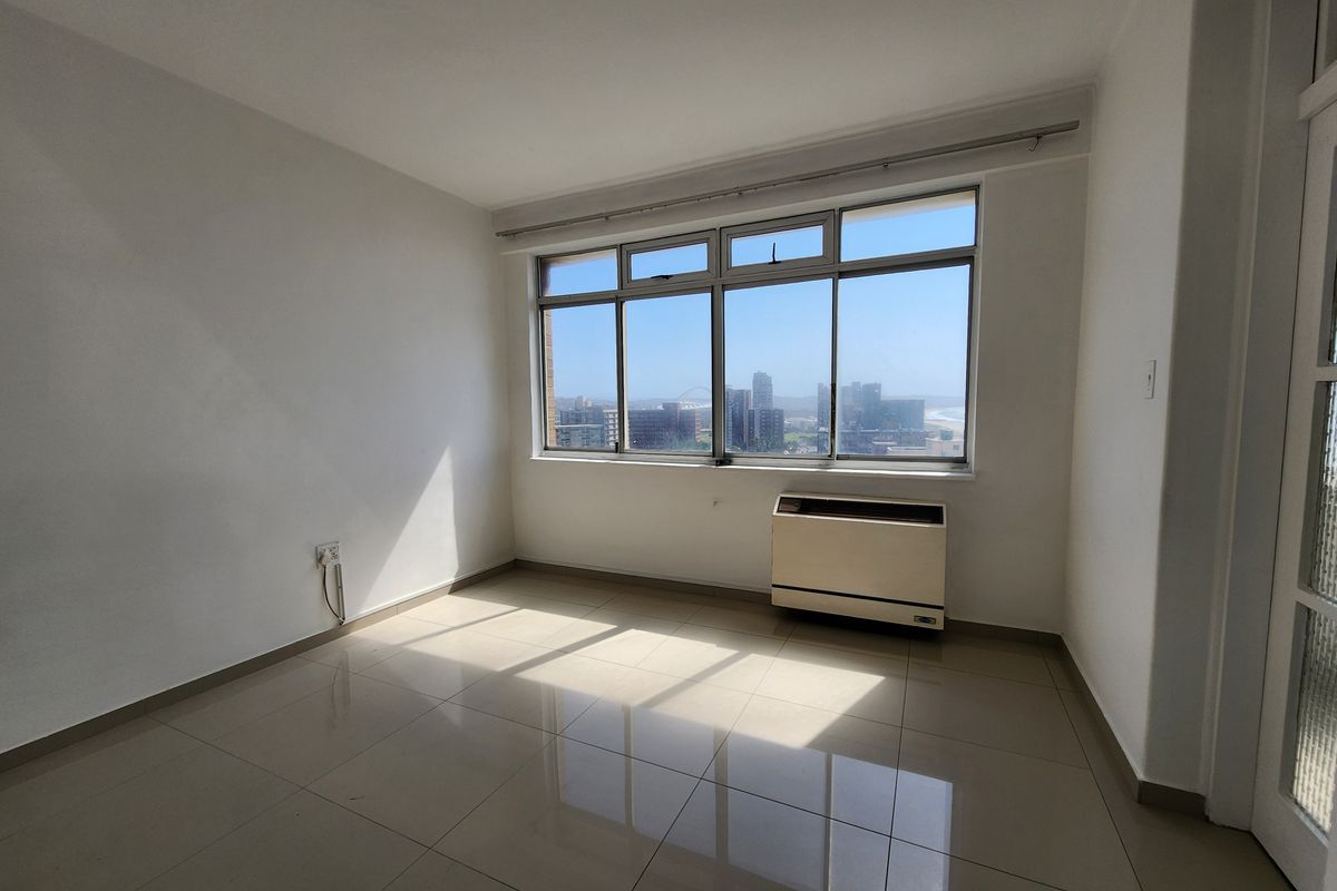 A bright, empty bedroom featuring neutral tiled flooring, large aluminium windows with city views, and a built-in air conditioning unit.