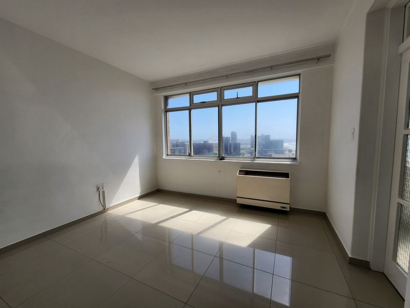 A bright, empty bedroom featuring neutral tiled flooring, large aluminium windows with city views, and a built-in air conditioning unit.