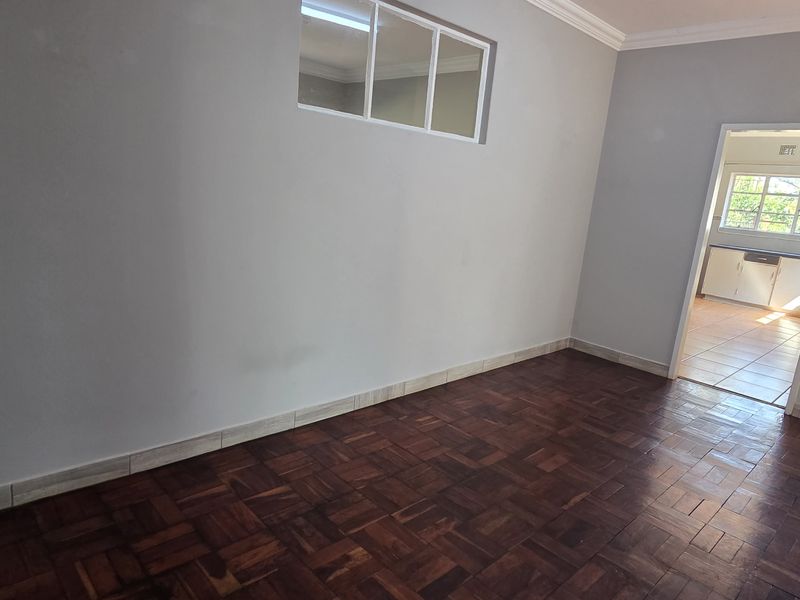 An empty living room featuring classic parquet flooring, neutral walls, and a view through to the kitchen area.