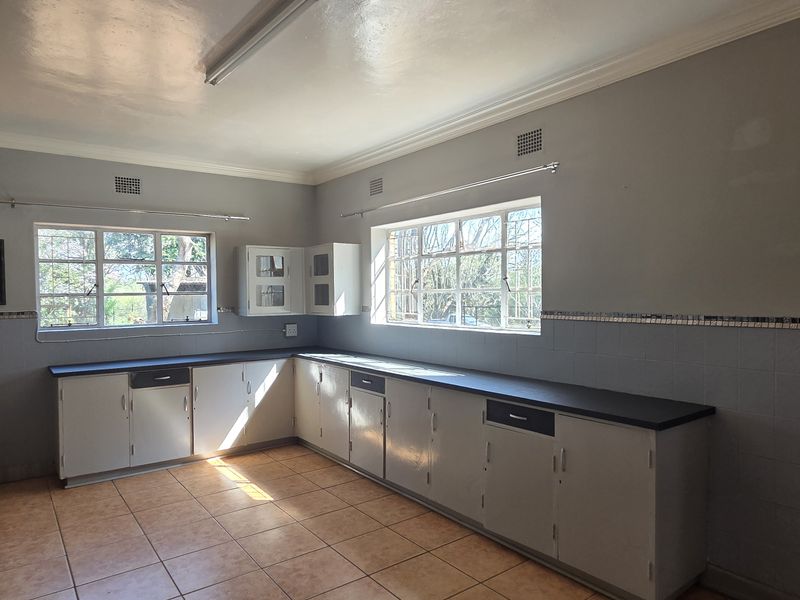 A spacious, empty kitchen featuring built-in cabinetry, dark countertops, tiled flooring, and large windows providing ample natural light.