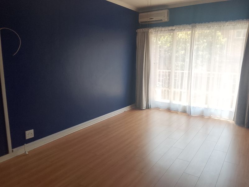 An empty living room featuring laminate flooring, a bold blue accent wall, and a wall-mounted air conditioning unit with access to a balcony.