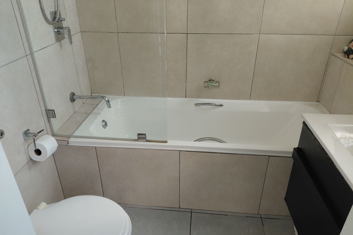 A modern bathroom featuring a built-in bathtub with a glass shower screen, wall-mounted fixtures, a toilet, and a vanity cabinet with neutral tiling.