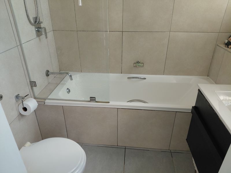 A modern bathroom featuring a built-in bathtub with a glass shower screen, wall-mounted fixtures, a toilet, and a vanity cabinet with neutral tiling.