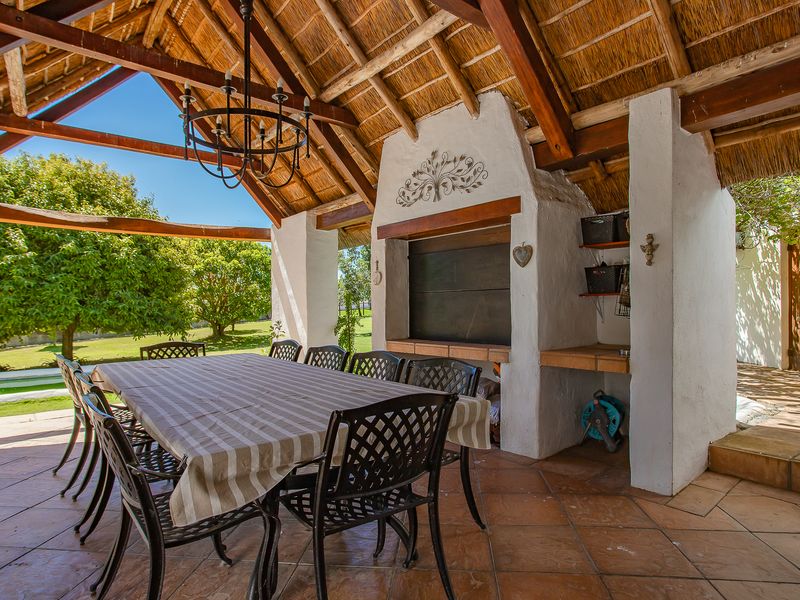 Spacious covered patio featuring a traditional thatched roof, exposed timber trusses, and a built-in braai, perfect for outdoor dining and entertaining.