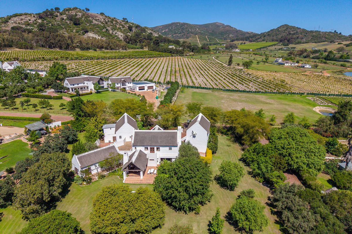 Aerial view of a large estate featuring multiple white buildings with thatched roofs, surrounded by expansive lawns, mature trees, and scenic vineyard landscapes.