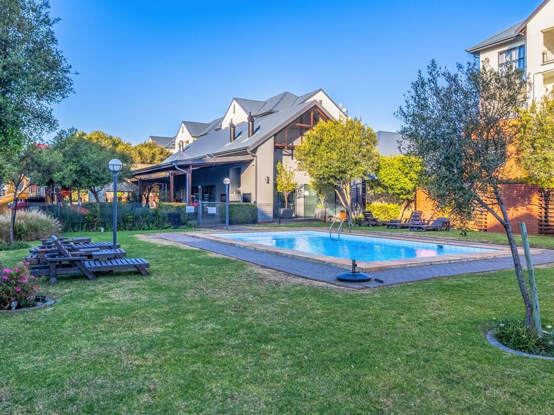 A well-maintained communal swimming pool area featuring a paved surround, landscaped lawn, and established trees within a townhouse complex.