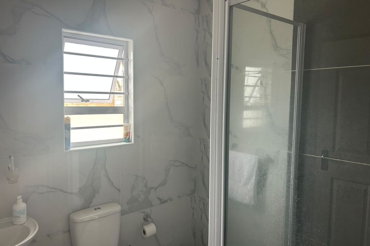 A modern bathroom featuring marble-look wall tiles, a glass shower enclosure, a wall-mounted basin, and a louvered window with security bars.