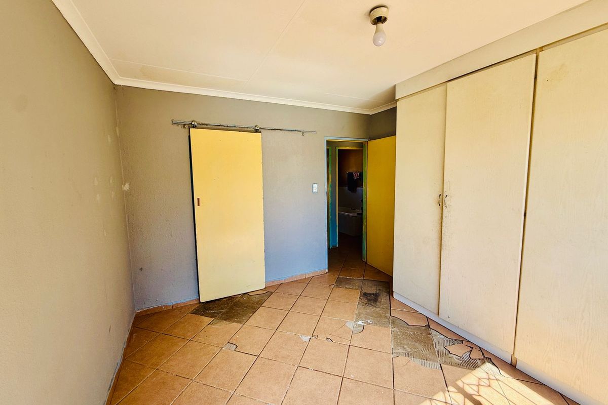 A bedroom featuring built-in cupboards, tiled flooring with visible damage, and a sliding door, requiring cosmetic updates and floor repairs.