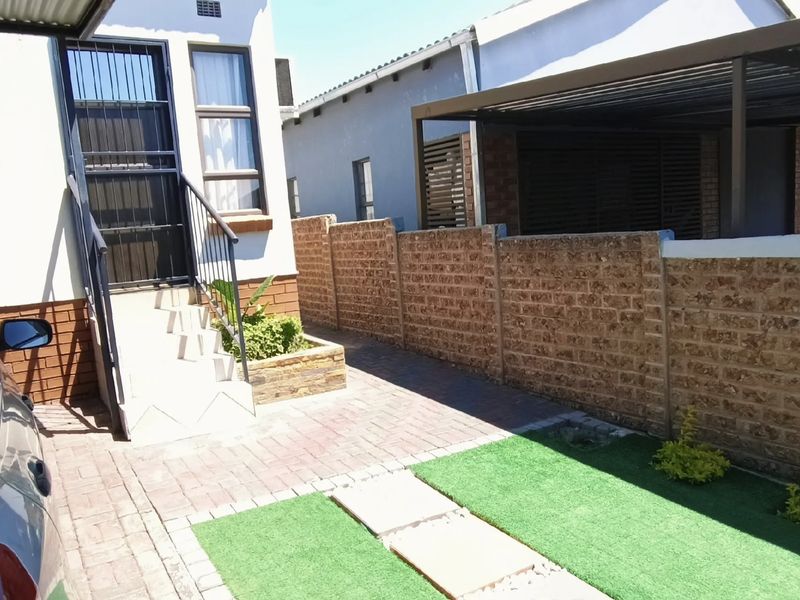 Front exterior view featuring a paved driveway, carport, brick boundary wall, and a small landscaped area with artificial grass leading to the entrance.