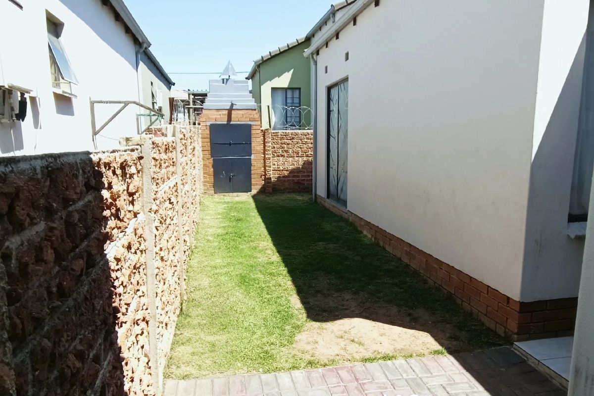 A narrow rear garden area featuring a lawn, paved walkway, and a built-in brick braai structure enclosed by boundary walls.