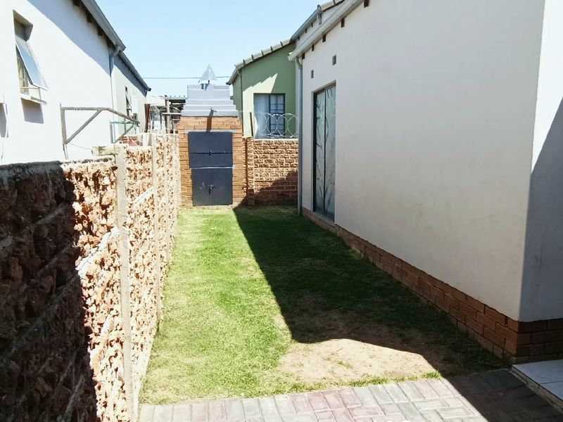 A narrow rear garden area featuring a lawn, paved walkway, and a built-in brick braai structure enclosed by boundary walls.