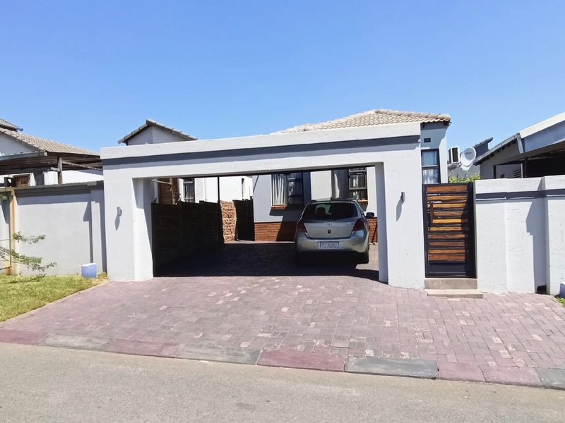 Front view of the property featuring a paved driveway, a covered carport, and secure boundary walling with a pedestrian gate.