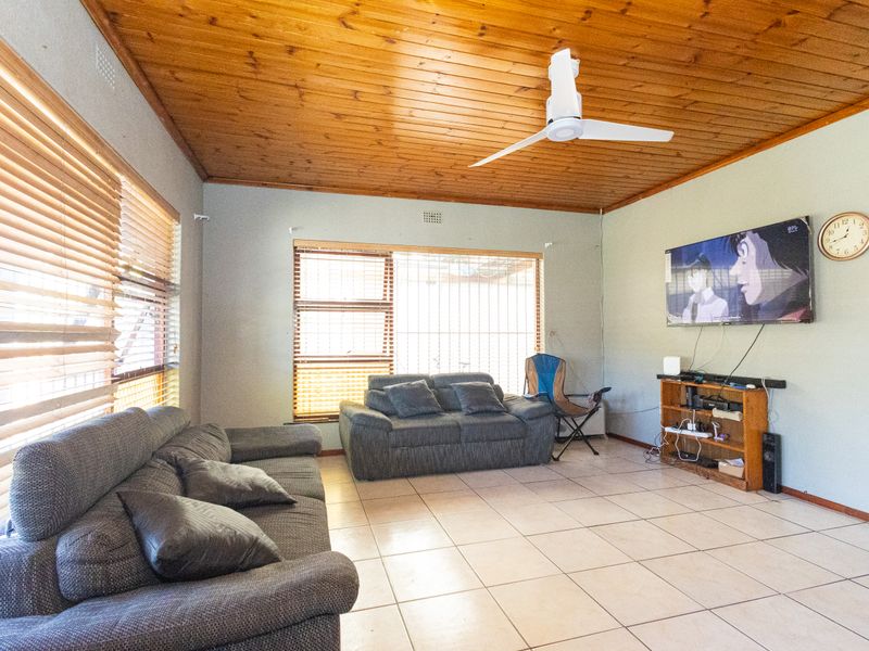A living room featuring a timber tongue and groove ceiling, tiled flooring, and large windows with venetian blinds.