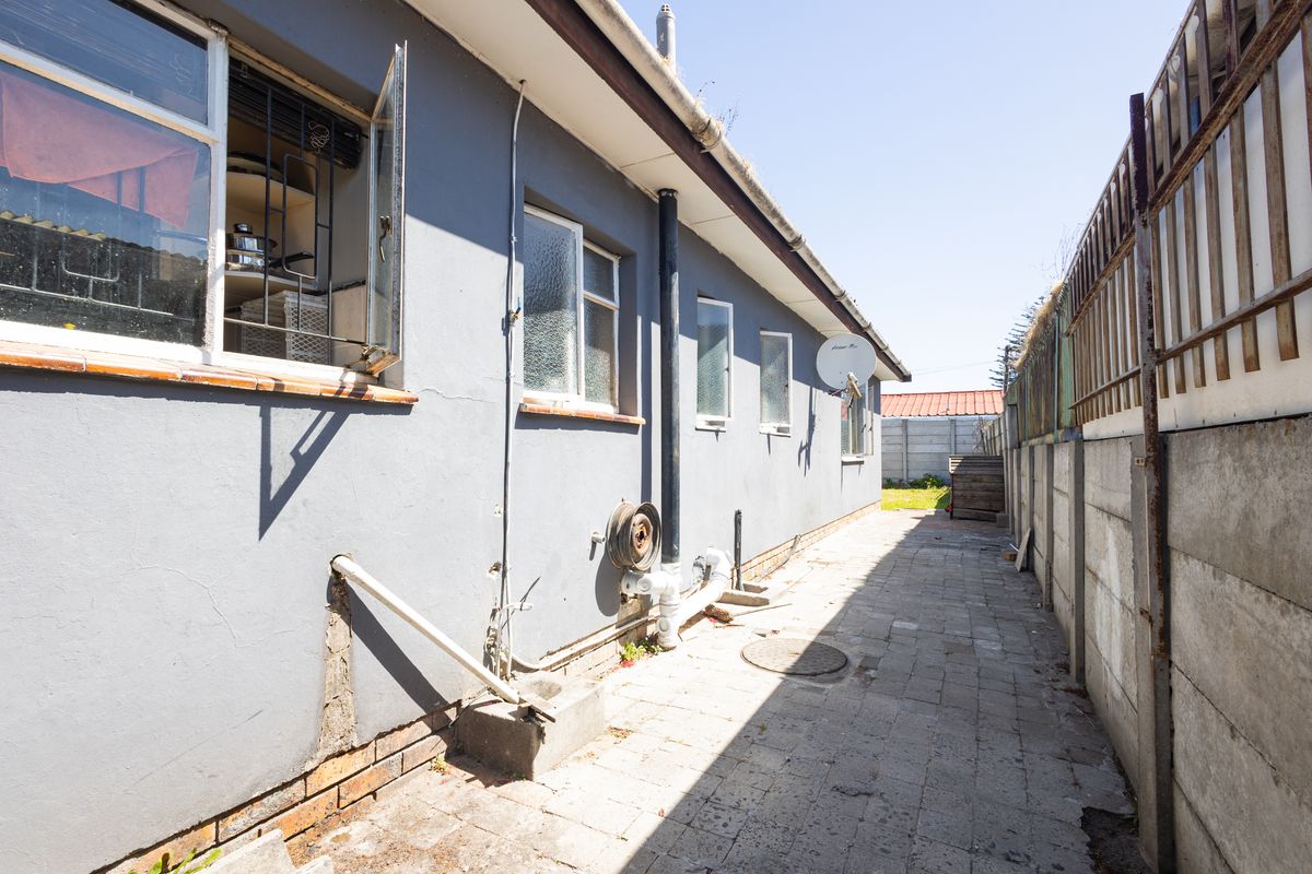 A narrow rear exterior walkway featuring paved flooring, precast concrete boundary walls, and visible external plumbing along the side of the house.