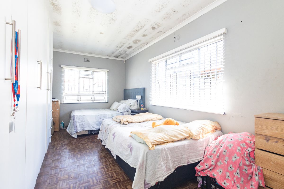 Spacious bedroom featuring parquet flooring, built-in cupboards, and large windows with blinds. The ceiling shows significant signs of water damage requiring repair.