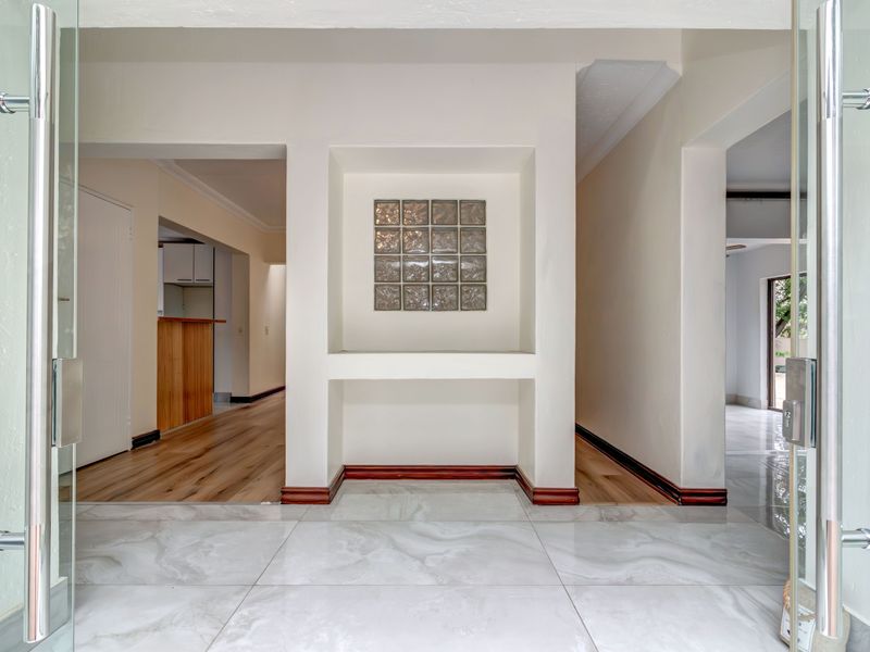 A bright and welcoming entrance hall featuring a modern glass pivot door, elegant marble-look floor tiles, and a decorative built-in wall niche.
