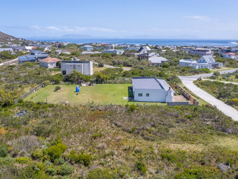 Aerial view of a residential plot featuring a grass lawn, fenced boundaries, and a single-story dwelling with a chimney, set within a coastal neighborhood.