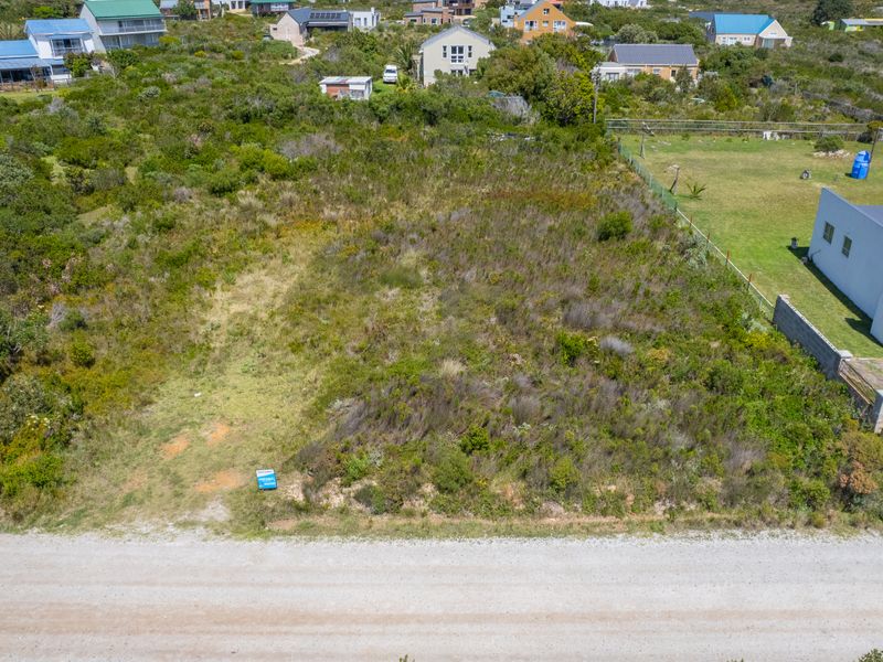 An aerial view of a vacant residential plot featuring natural vegetation, situated in a developed coastal neighborhood with existing perimeter fencing.