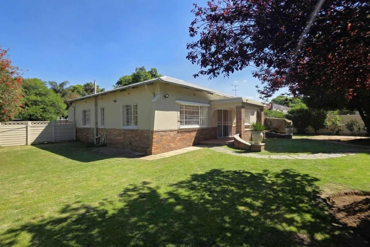 A view of the rear exterior of the house featuring a brick and plaster facade, security bars on windows, and a spacious garden with paved walkway.