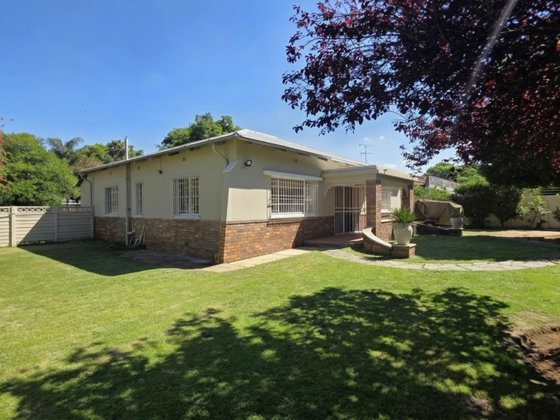 A view of the rear exterior of the house featuring a brick and plaster facade, security bars on windows, and a spacious garden with paved walkway.