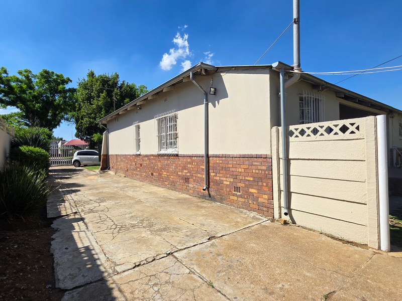 Rear exterior view of the house featuring a brick and plaster facade, security bars on windows, and a concrete driveway with visible surface cracking.