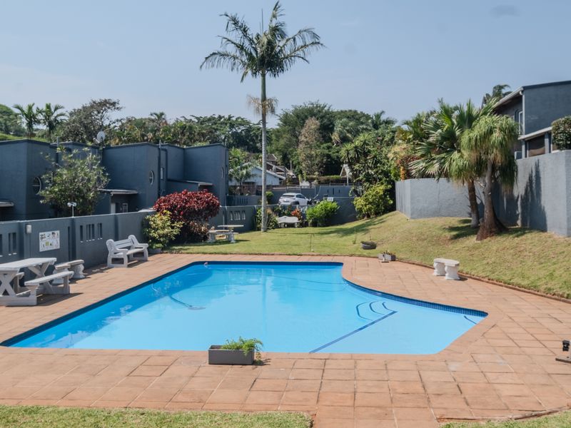 A well-maintained communal swimming pool area featuring a paved surround, landscaped garden, and secure boundary walls within the townhouse complex.